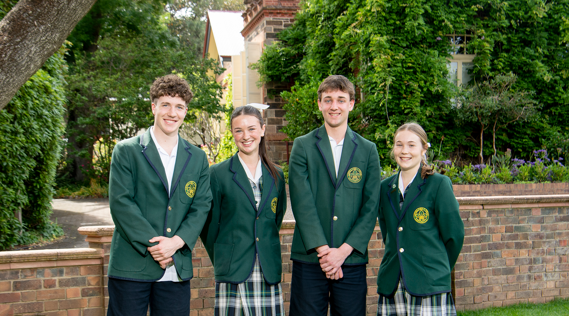 Group of two male and two female Mercedes College students in formal uniform standing in front of historic campus buildings.